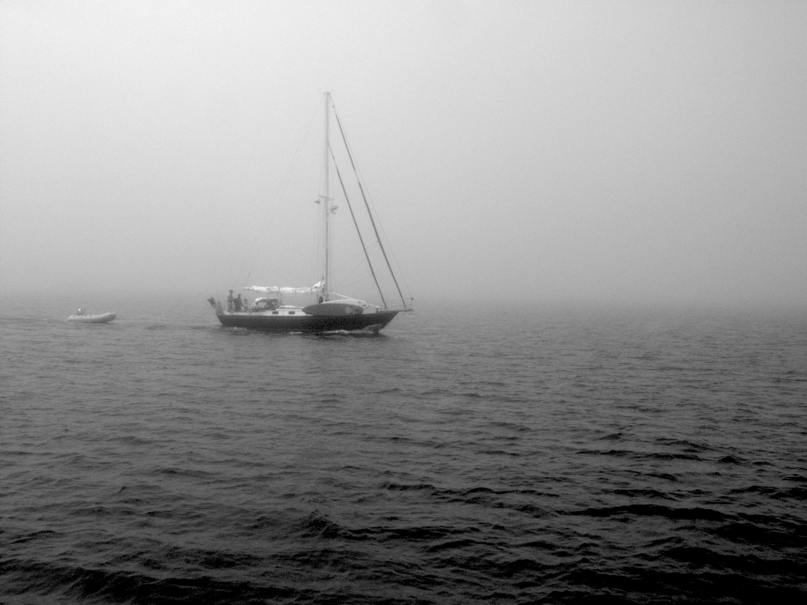 A black and white photo of a sailboat taxiing back into Boothbay Harbor, Maine in dense fog.